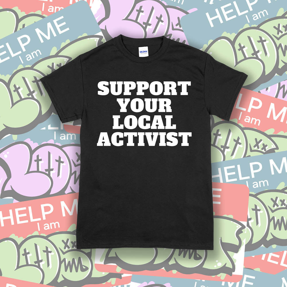 The original Support Your Local Activist t-shirt. Bold white letters screen printed on a black shirt. The shirt is displayed against a wall of colorful graffiti stickers.
