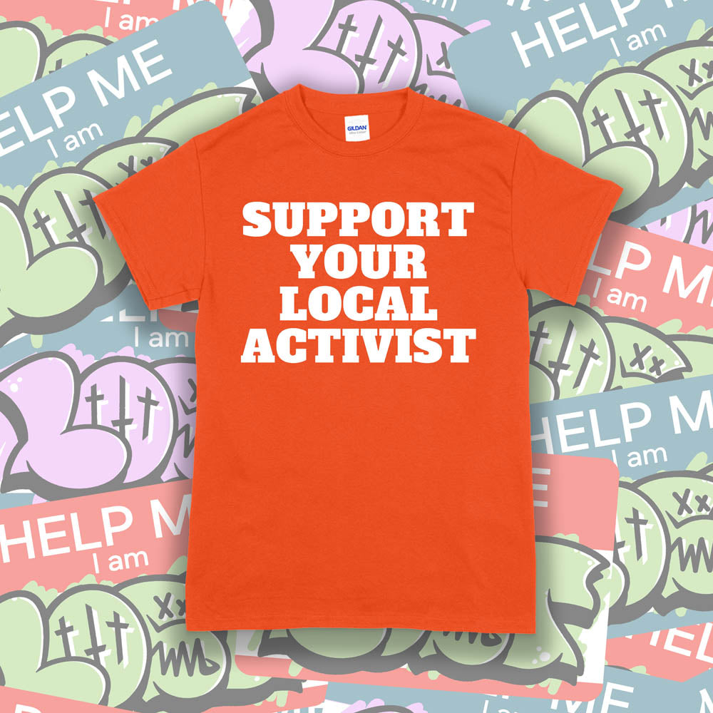 The original Support Your Local Activist t-shirt. Bold white letters screen printed on an orange shirt. The shirt is displayed against a wall of colorful graffiti stickers.