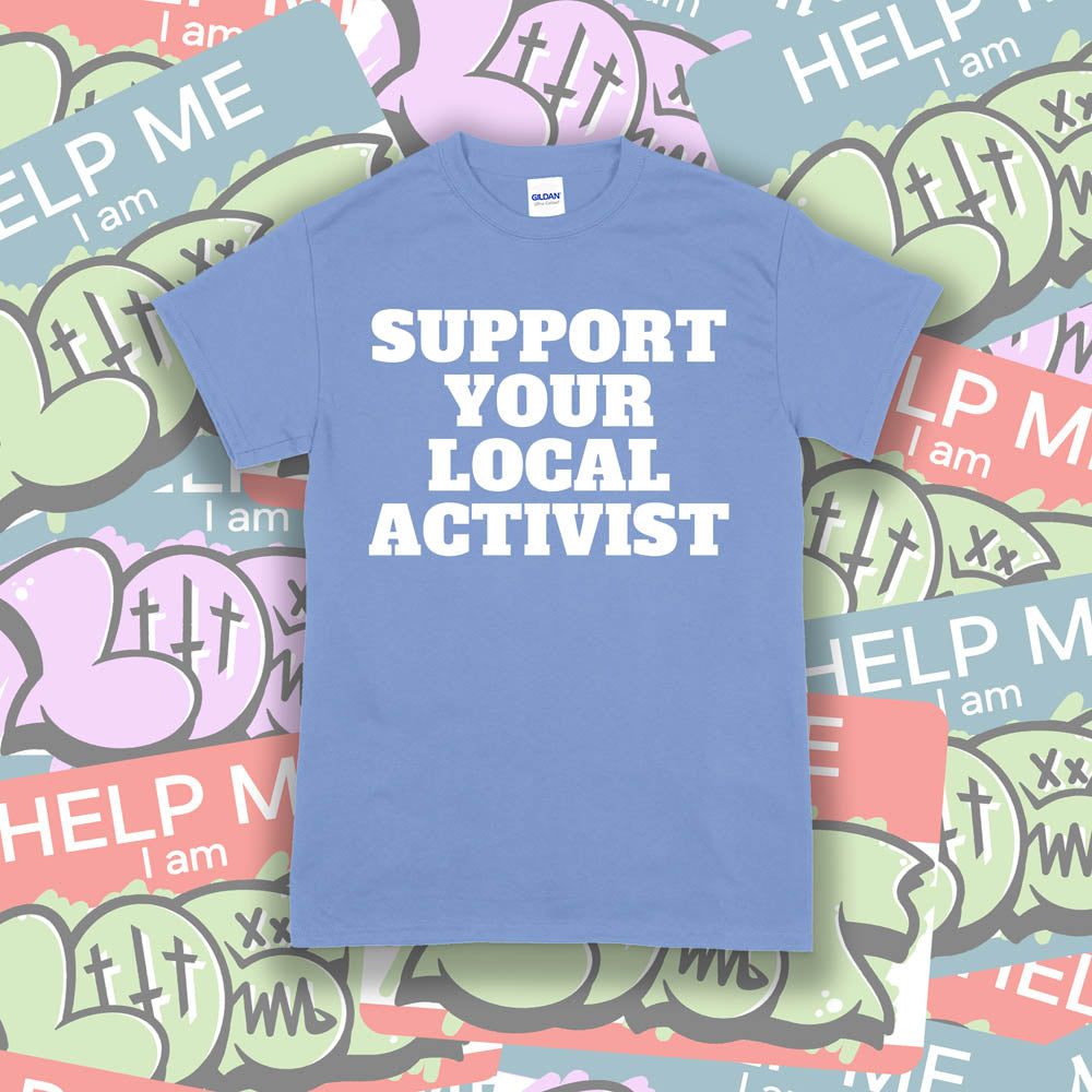 The original Support Your Local Activist t-shirt. Bold white letters screen printed on a light blue shirt. The shirt is displayed against a wall of colorful graffiti stickers.