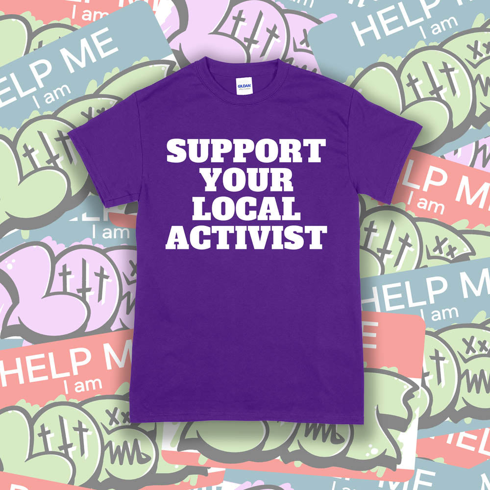 The original Support Your Local Activist t-shirt. Bold white letters screen printed on a purple shirt. The shirt is displayed against a wall of colorful graffiti stickers.