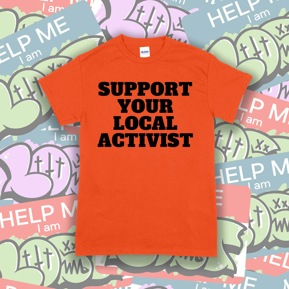 The original Support Your Local Activist t-shirt. Bold black letters screen printed on an orange shirt. The shirt is displayed against a wall of colorful graffiti stickers.
