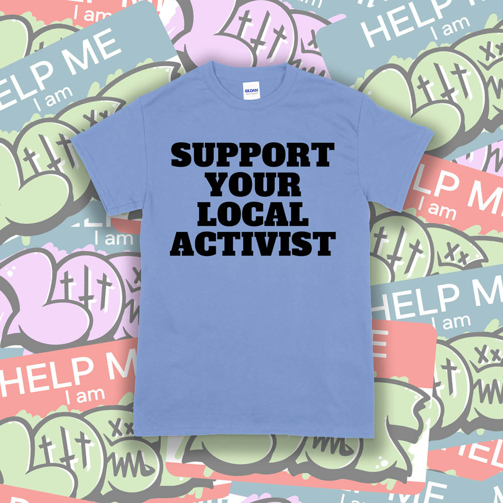 The original Support Your Local Activist t-shirt. Bold black letters screen printed on a light blue shirt. The shirt is displayed against a wall of colorful graffiti stickers.