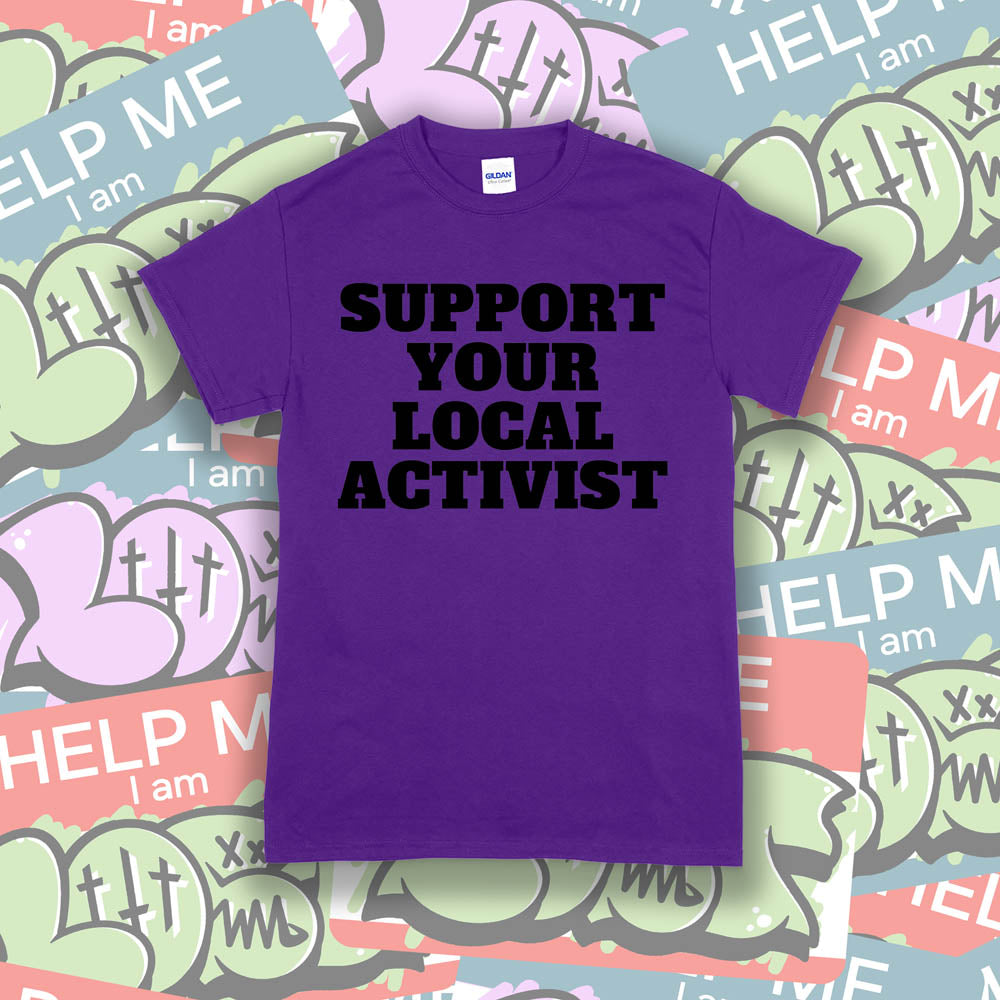 The original Support Your Local Activist t-shirt. Bold black letters screen printed on a purple shirt. The shirt is displayed against a wall of colorful graffiti stickers.